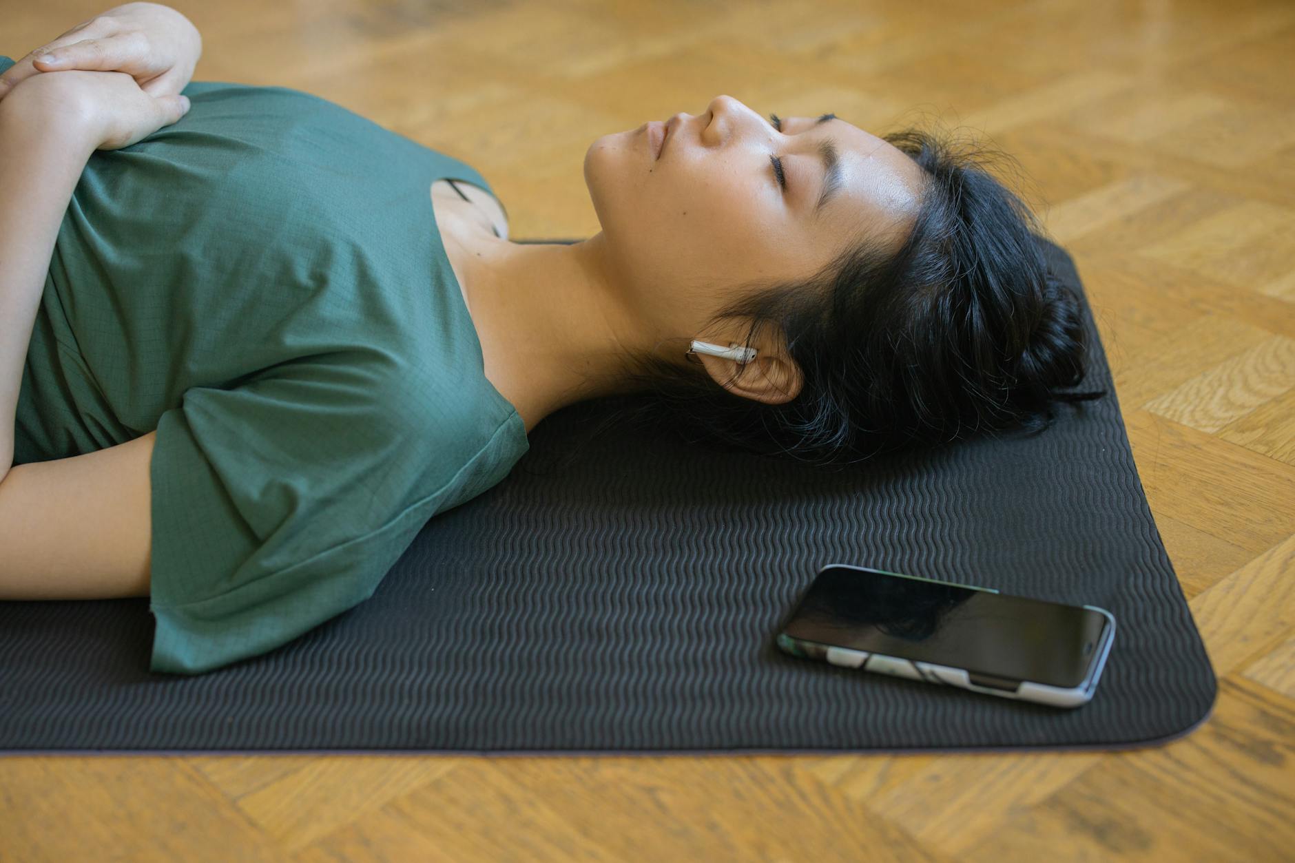 Asian woman resting on yoga mat with wireless earbuds and smartphone indoors.