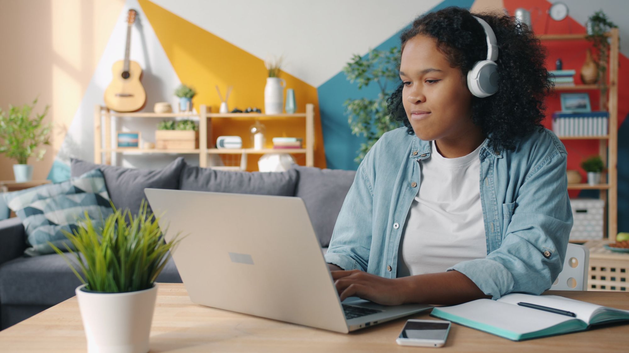 A woman wearing headphones looks at a laptop, with an open notebook and a pen on the desk