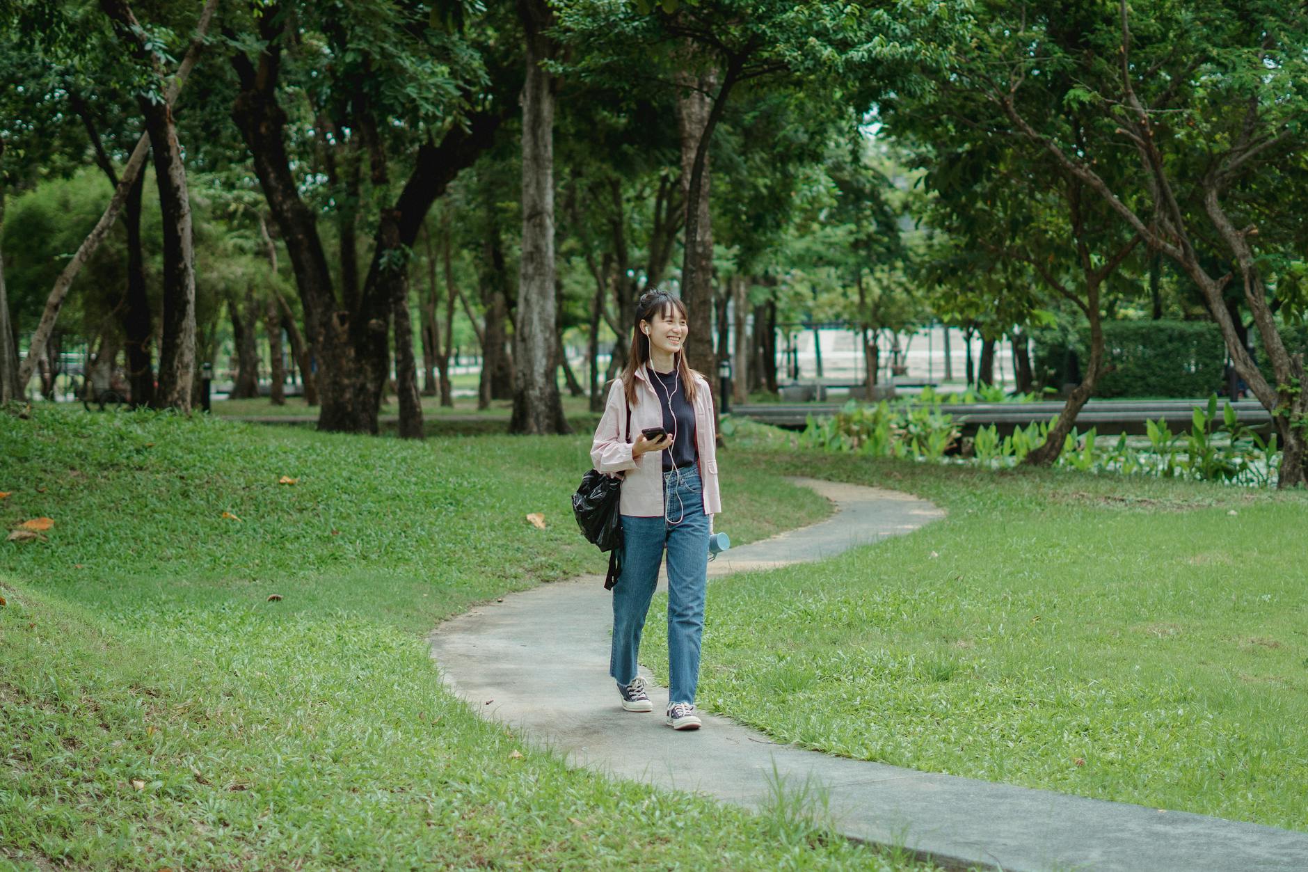 IMAGE: A person wearing earbuds and a backpack, walking through a park with lush greenery and dappled sunlight